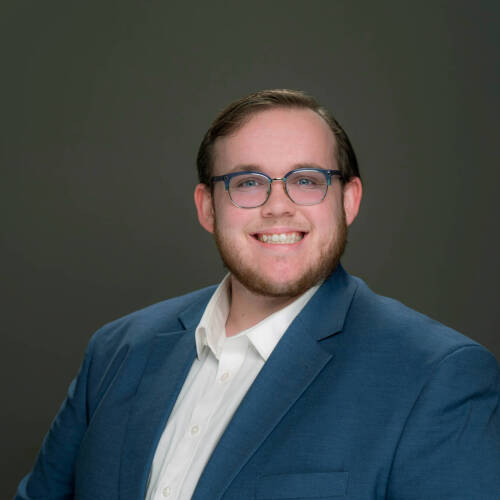 Ben Robar seated for portrait in blue jacket and white, open-collar shirt